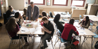 An educator stands at the head of a table filled with students in conversation.