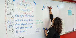 An educator writes with markers on a large piece of paper in a classroom. On another paper in large letters is "Universe of Obligation."