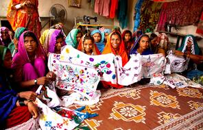 Young women seated on the floor of a room in colorful clothing and headscarves hold up a quilt they made.
