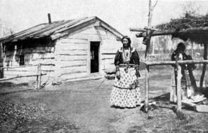 A woman stands in front of a small, run down wooden house. 