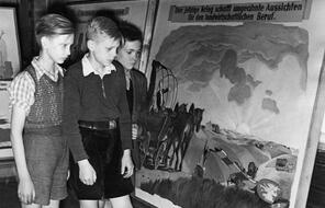 An exhibit at a Berlin school persuades Germans to help colonize the Warthegau area of Poland. The exhibit says “The land calls you!,” and the painting shows a settler’s car passing by a Polish border sign that has been knocked down.