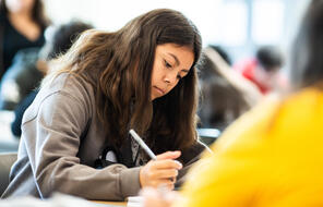 A student in a grey sweatshirt looks down at a paper with a pencil in hand.