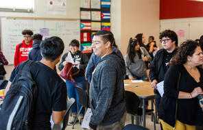 Students move around the classroom in conversation with each other. One student looks directly into the camera with a smile on their face.