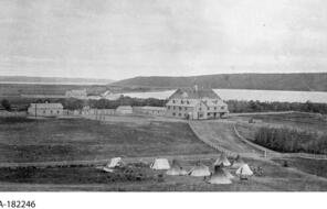 A rural setting with teepees in the foreground and a group of buildings in the background.