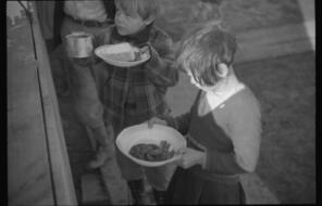 Two children hold plates with arms outstretched, seeking food.