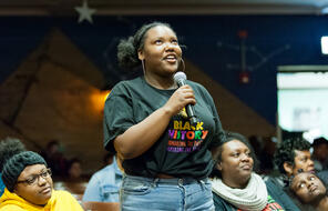 A Black student looks up while speaking into a microphone wearing a shirt that reads, "Black History: Honoring the Past, Inspiring the Future."