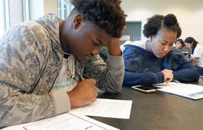 Two Black students write at their desks. 