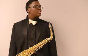 A Black teenager in a suit looks off in the distance while holding a saxophone.