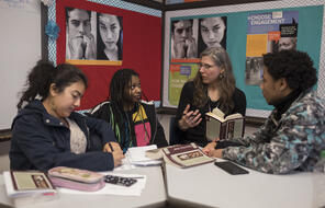 A sitting teacher speaks to three students while holding a book.