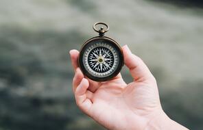 A person holds a compass in their hand with the ocean in the background.