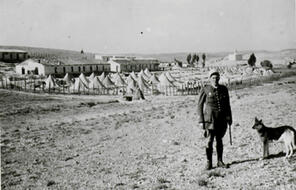A soldier stands in front of a camp with a dog.