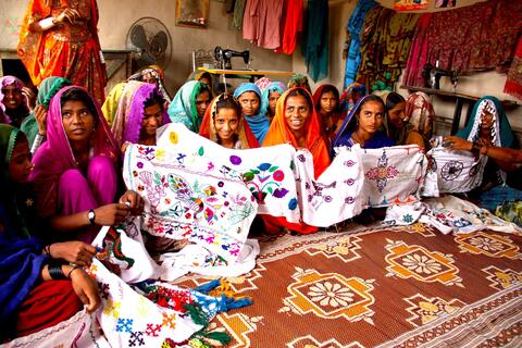 Young women seated on the floor of a room in colorful clothing and headscarves hold up a quilt they made.