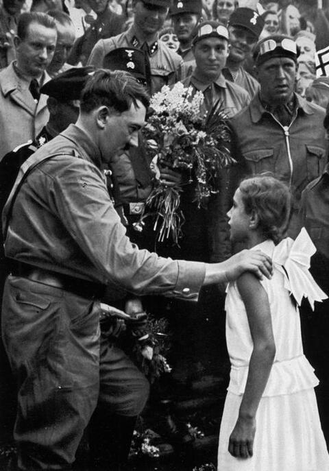Adolf Hitler has his hand placed on a young girl in a white dress as he speaks with her