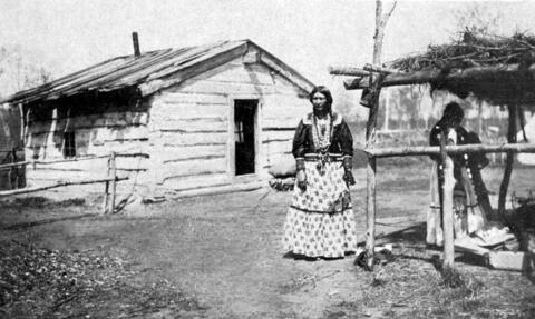 Une femme se tient devant une petite maison délabrée construite en bois. 