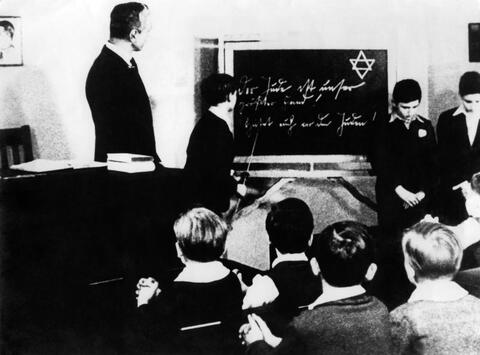 While this classroom studied “race science,” Jewish students were forced to stand at the front of the room. The blackboard says, “The Jew is our greatest enemy.”