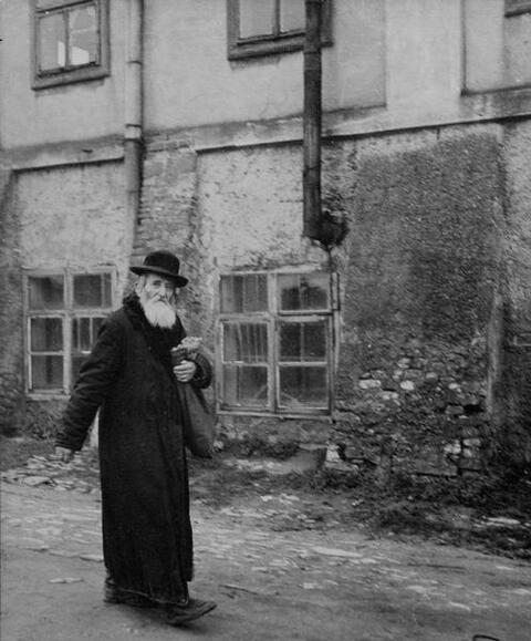 An elderly man with a long white beard, wearing a black hat and a long black coat walks down a street carrying a sack.