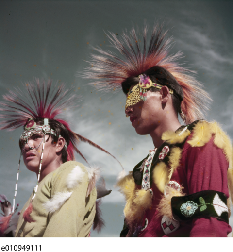 Two men wearing feathered and beaded headwear and clothing celebrate the sundance.