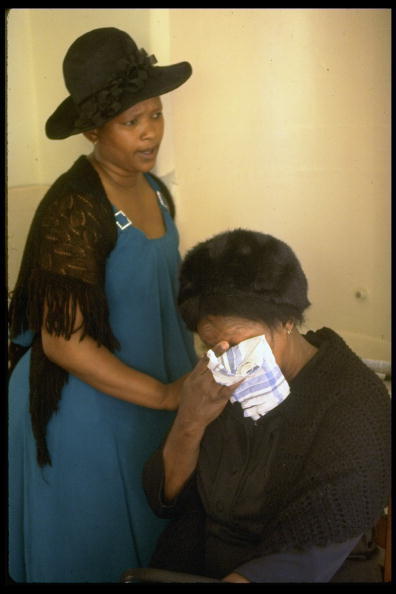 Two black South African women. Younger woman is standing, wearing a blue dress and black shawl and hat, and is consoling the sitting older woman who is crying into a handkerchief