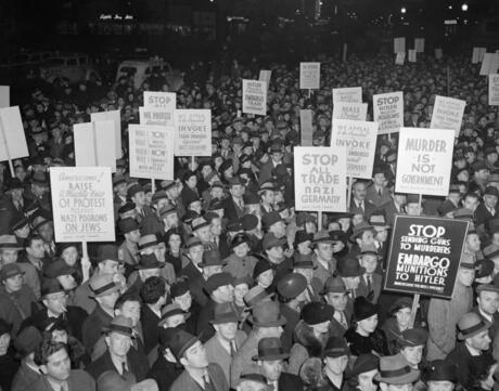 A crowd of American men and women hold signs protesting Nazi Germany's actions.