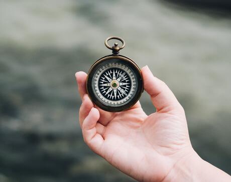 A person holds a compass in their hand with the ocean in the background.